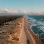 Vue aérienne d'une longue plage de sable bordée par l'océan avec des vagues, une forêt dense à gauche et un ciel nuageux