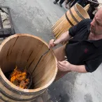 Un homme en polo noir tient un grand tonneau en bois avec un feu ardent à l'intérieur, lors d'une démonstration traditionnelle à Bordeaux.