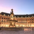 Place de la Bourse à Bordeaux illuminée la nuit, avec ses bâtiments historiques en pierre de taille, une fontaine centrale et des personnes se promenant sur la place pavée.