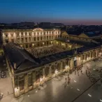 Vue nocturne de la Place de la Bourse à Bordeaux avec ses bâtiments historiques illuminés en jaune doré et des personnes circulant dans la place pavée.