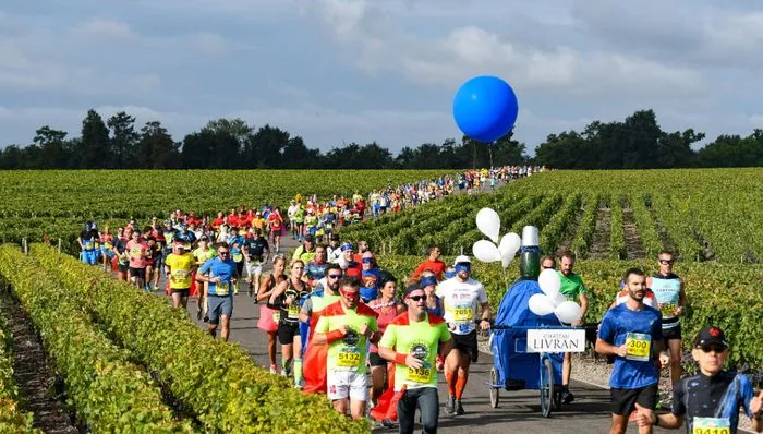 Groupe de coureurs participant au Marathon des Châteaux du Médoc traversant un vignoble bordelais, avec un ballon bleu géant et des pancartes de soutien.