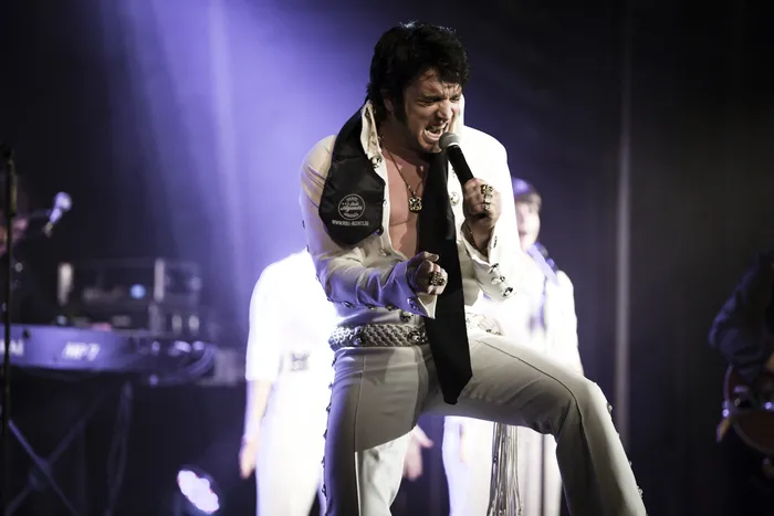 Performer en costume blanc et gilet noir avec ceinture ornée sur scène, microphone en main, dans une mise en scène évoquant l'univers d'Elvis lors du spectacle The Musical Story of Elvis à Bordeaux.