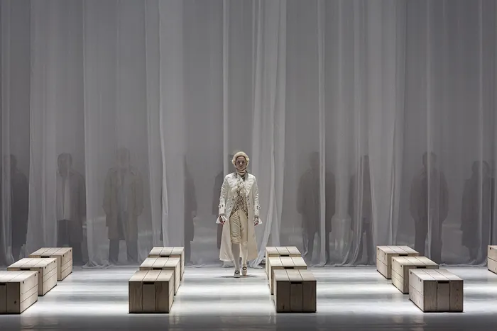 Sur une scène minimaliste avec des blocs de bois et un rideau gris striché, un chanteur en costume clair et chapeau se tient au centre, entouré de silhouettes floues en arrière-plan, dans une mise en scène du Requiem de Mozart à Bordeaux.