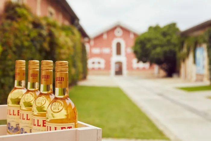Quatre bouteilles de Lillet alignées sur un présentoir blanc devant le château historique de la Maison Lillet à Bordeaux, avec un parc verdoyant et des bâtiments en arrière-plan