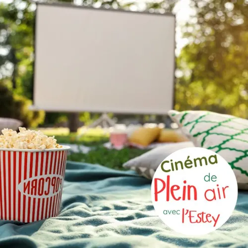 Scène de cinéma en plein air avec un seau de pop-corn à rayures rouges et blanches au premier plan, un écran de projection flou en arrière-plan, des coussins colorés sur la pelouse et des arbres verts, dans le cadre du Cinéma de plein air avec l'Estey à B