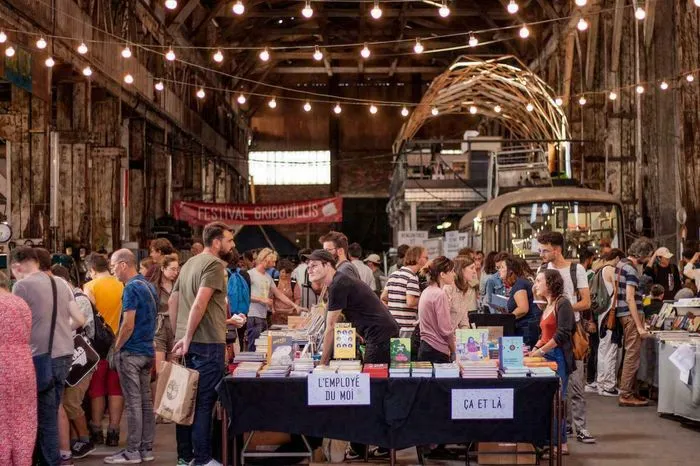 Foule de visiteurs lors du Festival Gribouillis à Bordeaux, avec des stands de livres et de produits artisanaux dans un entrepôt industriel illuminé par des guirlandes lumineuses.