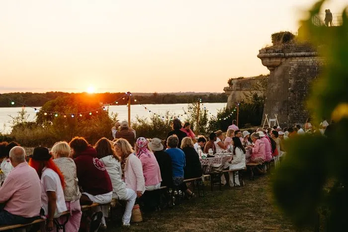 Dîner en plein air au coucher du soleil près de la Citadelle de Blaye avec des convives assis à des tables longues, face à l'eau et aux fortifications historiques.