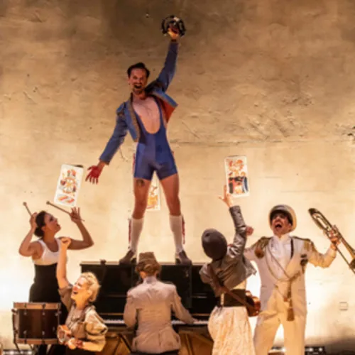 Scène de théâtre de l'opéra Carmen à Bordeaux : un danseur en costume bleu lève le poing au-dessus de sa tête, entouré de musiciens et de figurants jouant des instruments et tenant des affiches, sur fond de décor épuré.