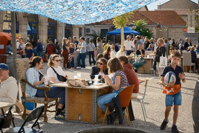 Foule de personnes dans une cour ensoleillée avec un plafond bleu ajouré, tables et chaises dispersées, enfants jouant, bâtiments en pierre en arrière-plan.