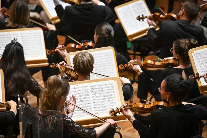 Vue d'ensemble d'un orchestre symphonique pendant une répétition ou un concert, avec des musiciens en noir tenant des violons et des partitions musicales sur des pupitres.