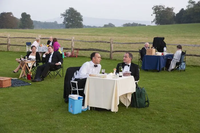 Pique-nique élégant en plein air dans un pré avec des convives en tenue formelle assis à des tables dressées, entourés de paysage rural et de clôtures en bois