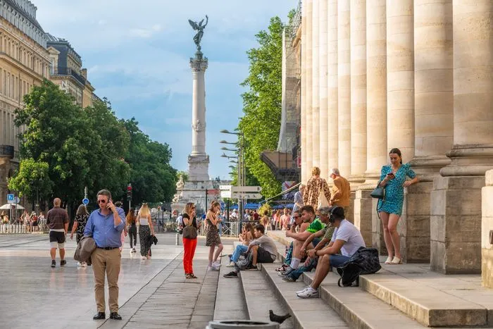 Place de la Bourse à Bordeaux avec ses colonnes majestueuses, des visiteurs assis sur les marches, et le monument aux Girondins visible en arrière-plan sous un ciel bleu.
