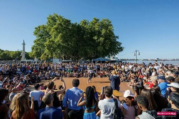 Grande foule de personnes rassemblées sur les quais de Bordeaux pour un événement festif de danse en plein air, avec des arbres, un monument et le port en arrière-plan sous un ciel bleu.