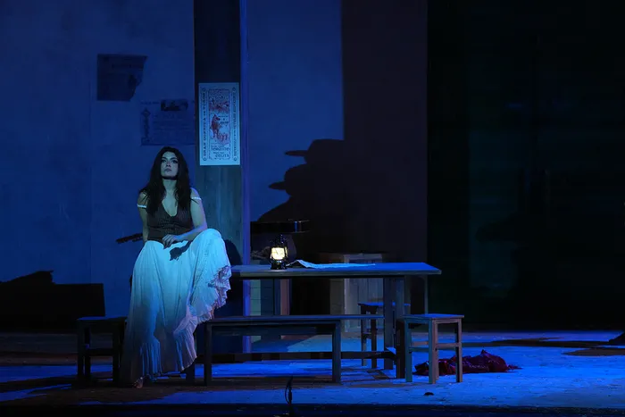 Scène de l'opéra Carmen au Théâtre National de Bordeaux : une femme en costume traditionnel assis à une table éclairée par des lumières bleues, dans un décor minimaliste avec des escaliers en arrière-plan.