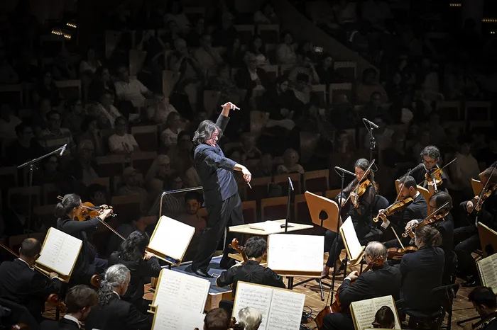 Un chef d'orchestre dirige un orchestre symphonique en concert, avec des musiciens jouant des instruments à cordes et à vent, partitions visibles, public en arrière-plan dans une salle de concert.