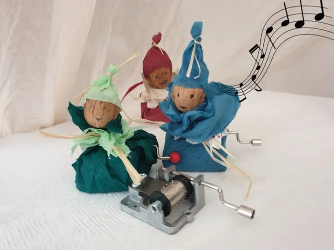 Boîte à musique artisanale avec trois personnages en tissu et papier mâché portant des chapeaux colorés (vert, rouge, bleu), montés sur un mécanisme à manivelle en métal, avec une portée musicale en arrière-plan