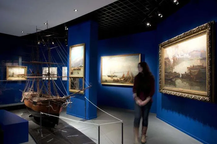 Galerie du Musée d'Aquitaine à Bordeaux avec murs bleu profond, maquette de navire ancien, peintures de marines historiques encadrées dorées, et visiteur observant les œuvres