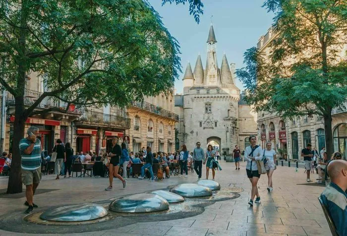 La Porte Cailhau, monument historique de Bordeaux, avec ses tours pointues caractéristiques, entourée d'une place publique animée avec des visiteurs et des arbres verts.