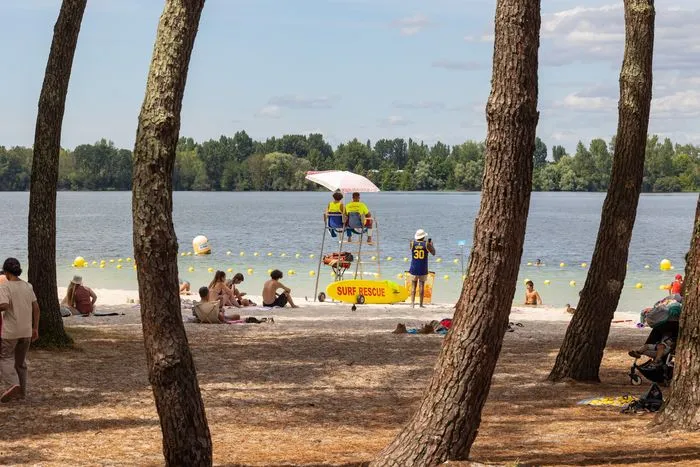 Plage du Lac à Bordeaux avec baigneurs, maîtres-nageurs en poste de surveillance jaune et blanc, parasol blanc, arbres côtiers, et zone de baignade délimitée par des bouées jaunes.