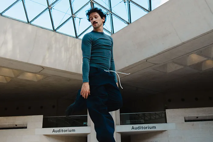 Homme posant dans un auditorium moderne de Bordeaux avec architecture contemporaine et verrière, vêtu d'un pull bleu pétrole et d'un pantalon bleu foncé