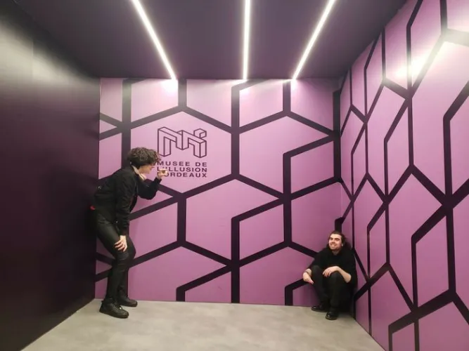 Deux visiteurs dans une salle du Musée de l'Illusion de Bordeaux, entourés de murs roses avec motifs géométriques noirs et éclairés par des néons blancs au plafond.