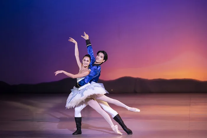 Deux danseurs de ballet en costume de prince et ballerine exécutent un pas de deux sur scène, avec un décor de coucher de soleil en arrière-plan.
