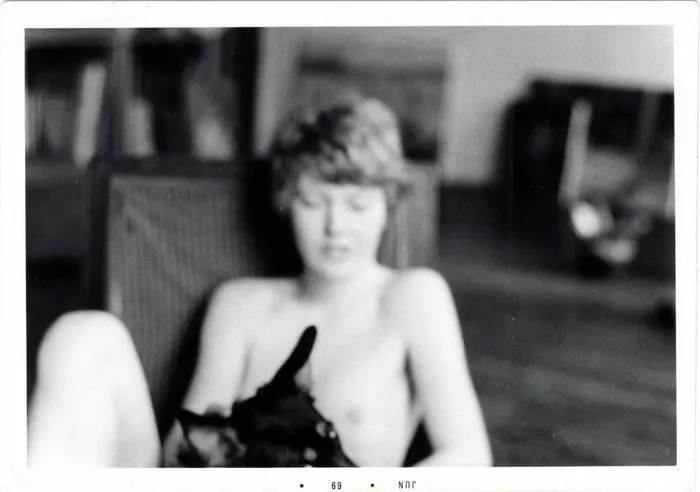 Photographie en noir et blanc d'une femme assise, vêtue d'un vêtement sans manches, dans un intérieur flou. Image d'archive numérisée datée de 1969.