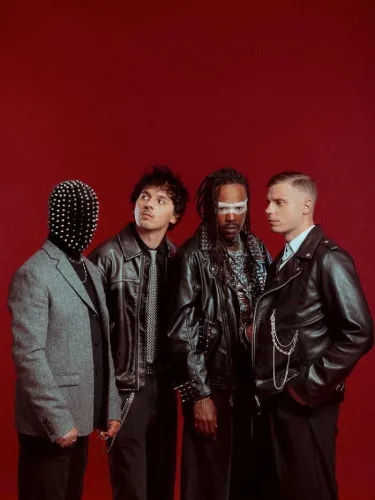 Quatre hommes posant face à la caméra sur un fond rouge uniforme. Le premier porte un masque à clous argentés et un blazer gris, le deuxième un cuir noir avec chemise à motifs, le troisième un cuir noir avec lunettes blanches et chaînes argentées, et le q