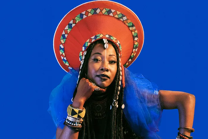 Portrait de Fatoumata Diawara portant un costume traditionnel avec un grand turban orange orné de motifs géométriques, sur fond bleu