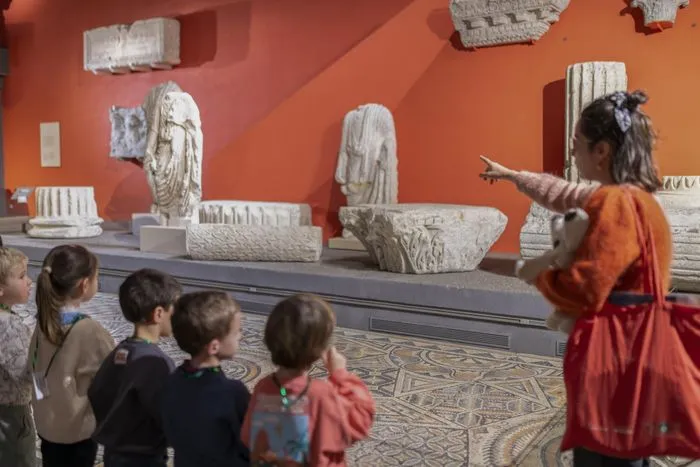 Une guide en costume orange explique à un groupe d'enfants des sculptures romaines exposées dans un musée à Bordeaux, avec des fragments de statues et d'objets antiques sur un mur orange.