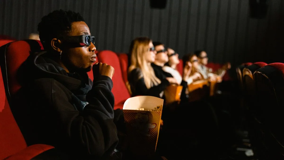Un spectateur portant des lunettes 3D regarde un film dans une salle de cinéma avec des fauteuils rouges, tenant un gobelet de pop-corn.
