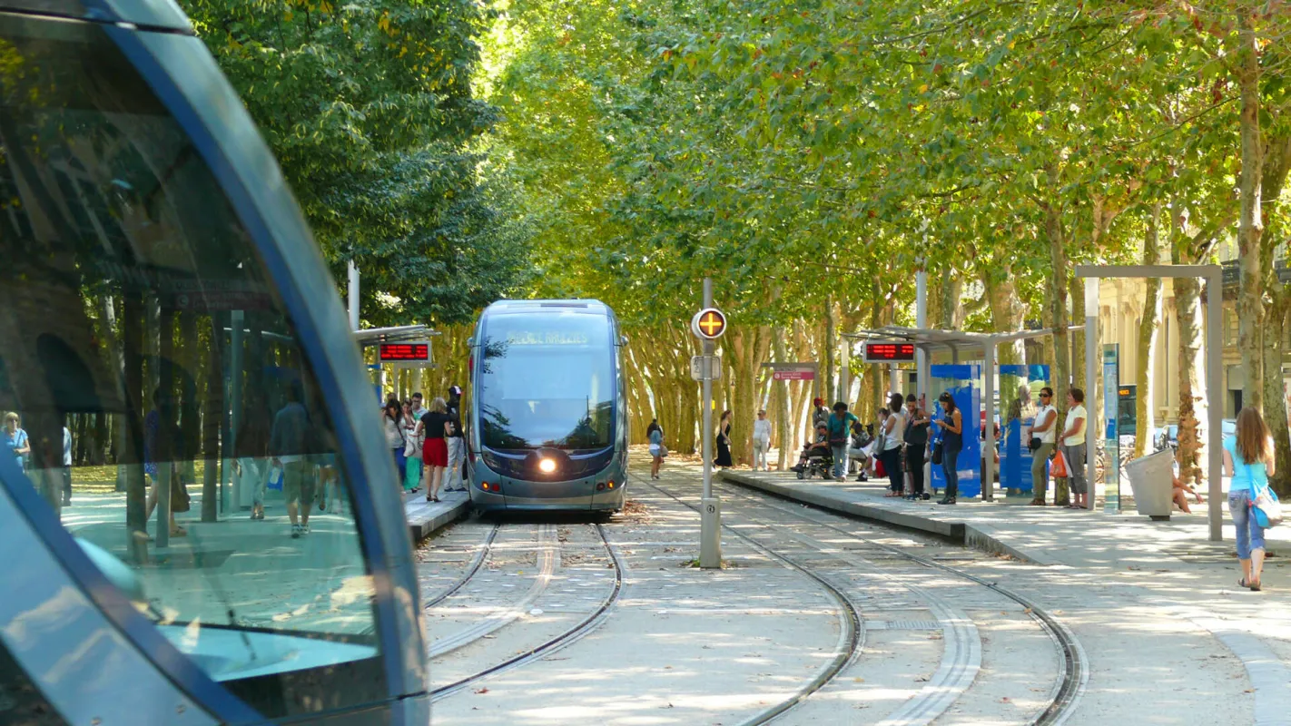 Station de tramway à Bordeaux avec un tramway gris en approche sur les rails, entouré d'arbres et de passagers attendant à la station