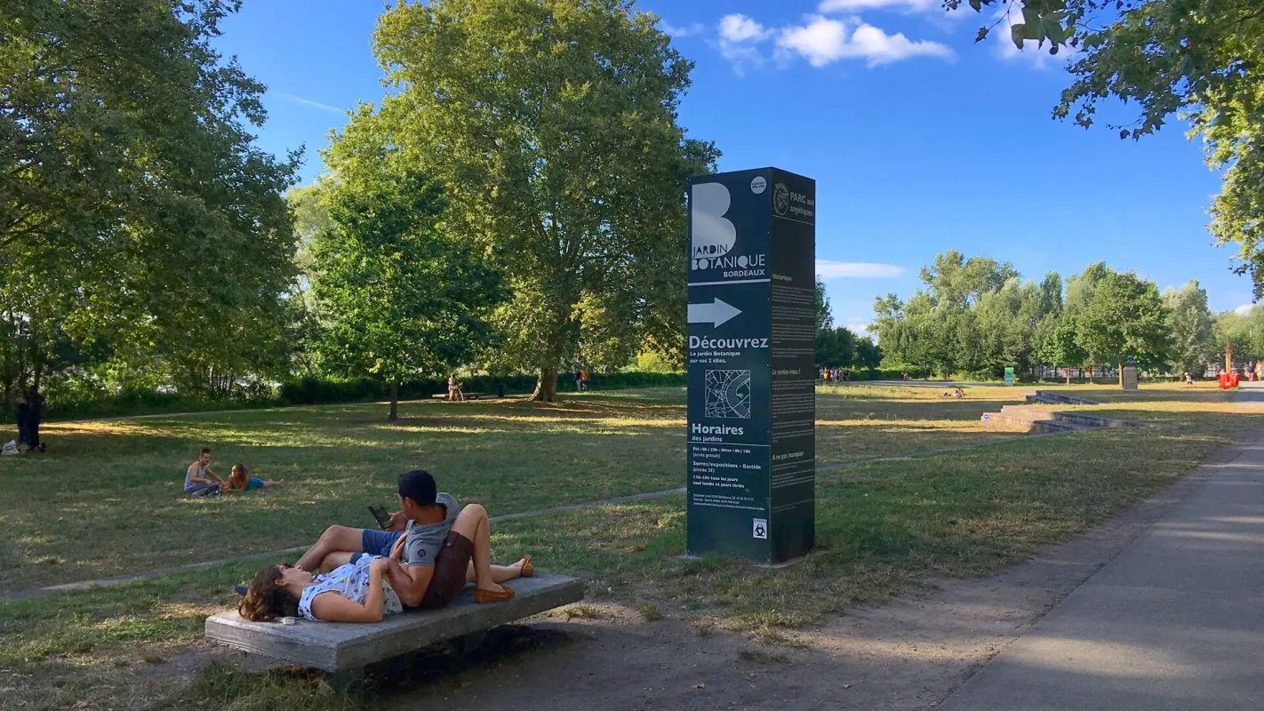 Famille allongée sur une plate-forme en béton dans le jardin botanique de Bordeaux par une journée ensoleillée, avec des arbres verts et un panneau d'information du parc visible.