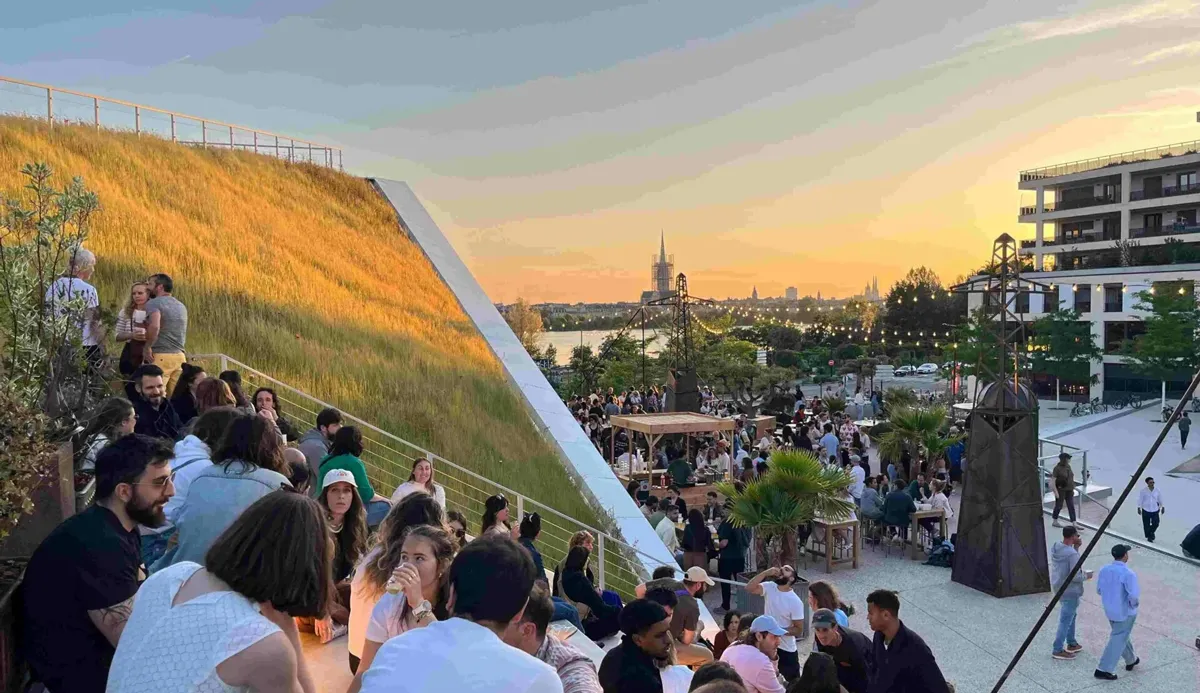 Vue panoramique d'une zone urbaine de Bordeaux avec une terrasse en pente couverte de végétation, des visiteurs profitant du coucher de soleil, et la skyline de la ville avec ses églises historiques en arrière-plan.