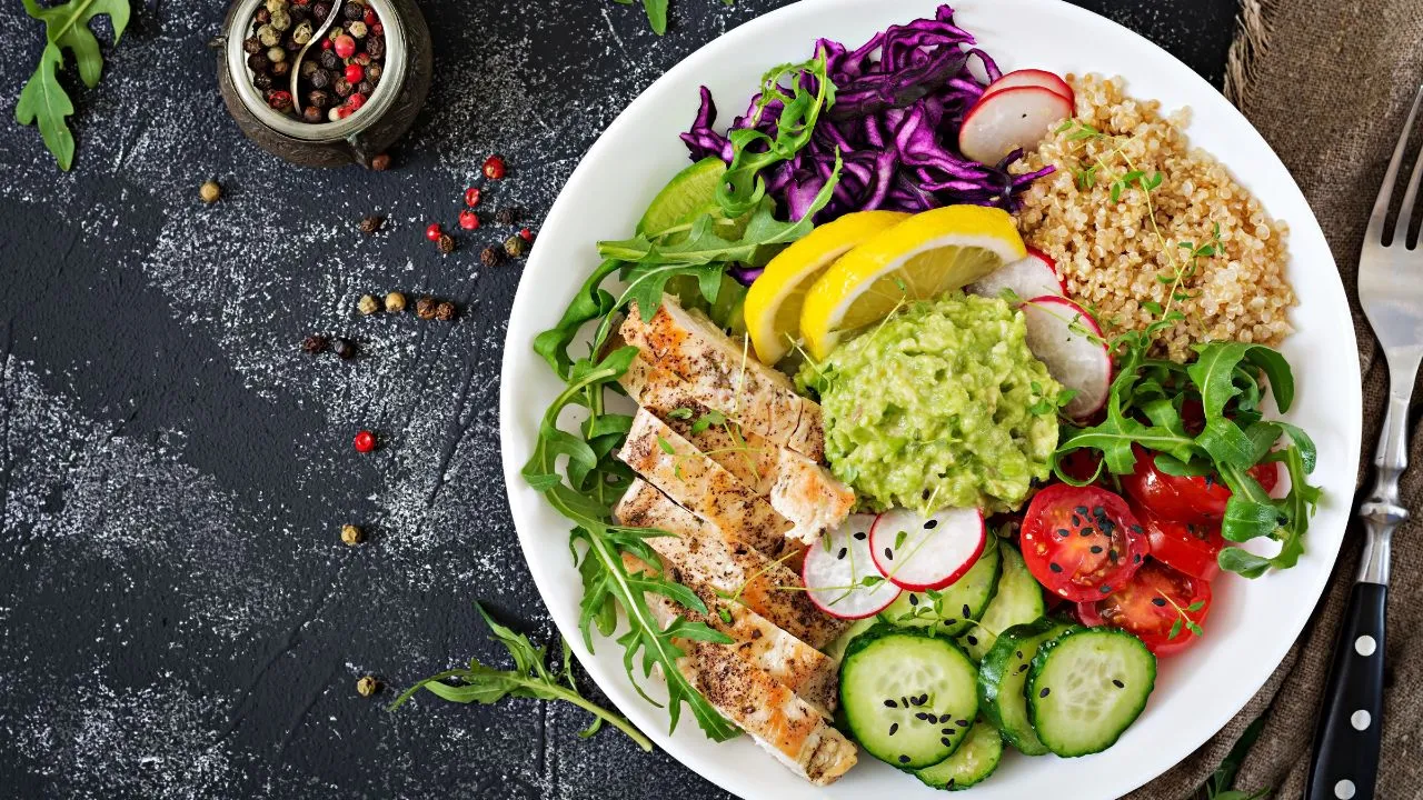 Assiette healthy avec filet de poulet grillé, quinoa, guacamole, légumes frais (tomates, concombre, radis, chou rouge), roquette et citron sur fond noir