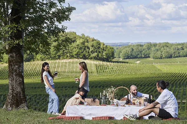 Un groupe de cinq personnes pique-nique sous un grand arbre à côté de vignobles à Bordeaux, avec une nappe blanche, des verres et des bouteilles de vin sur la table.