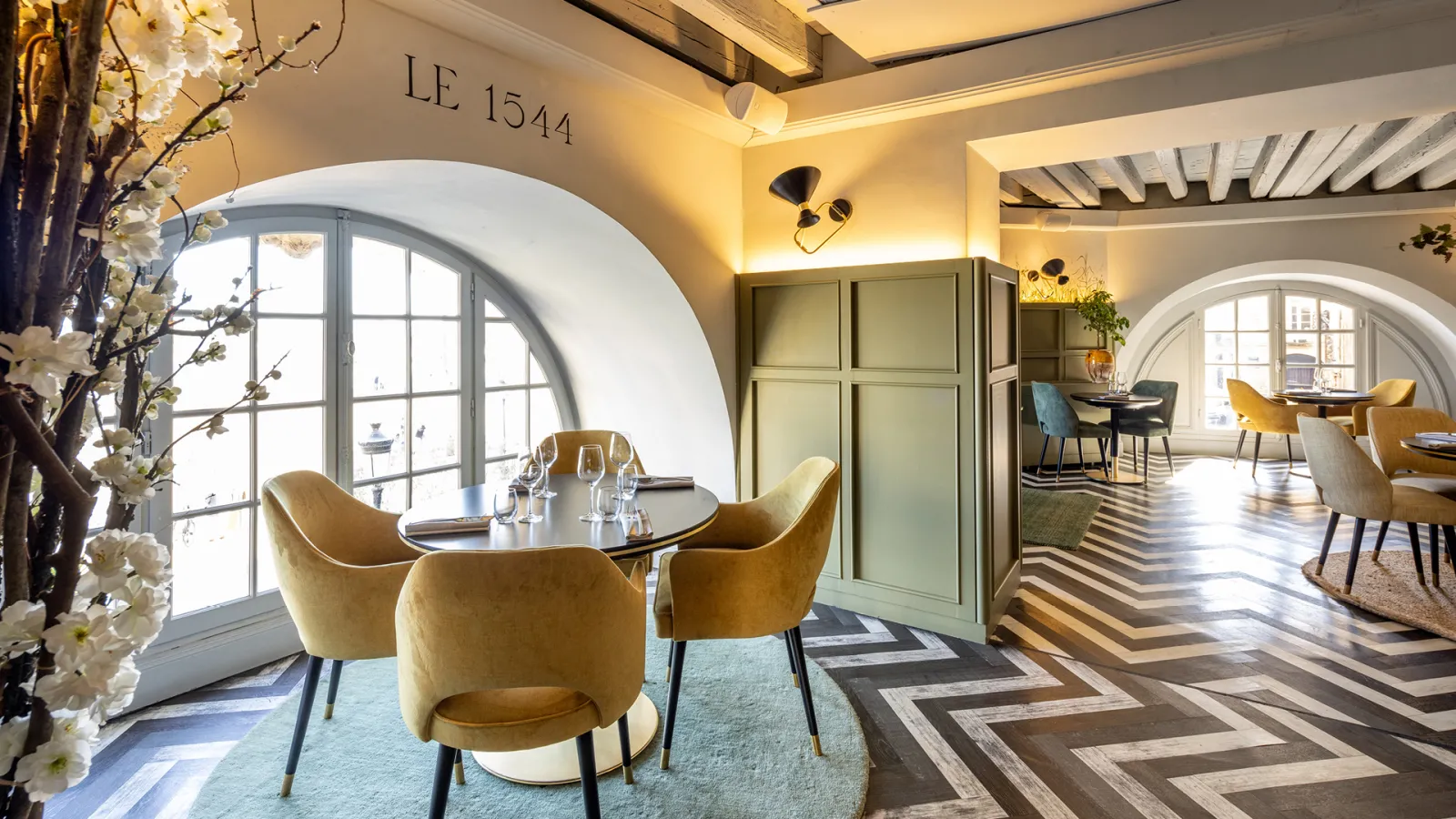 Salon élégant du Le 1544 à Bordeaux avec trois fauteuils dorés autour d'une table ronde, grande baie vitrée arquée avec fleurs blanches, plafond avec poutres apparentes et éclairage chaleureux.