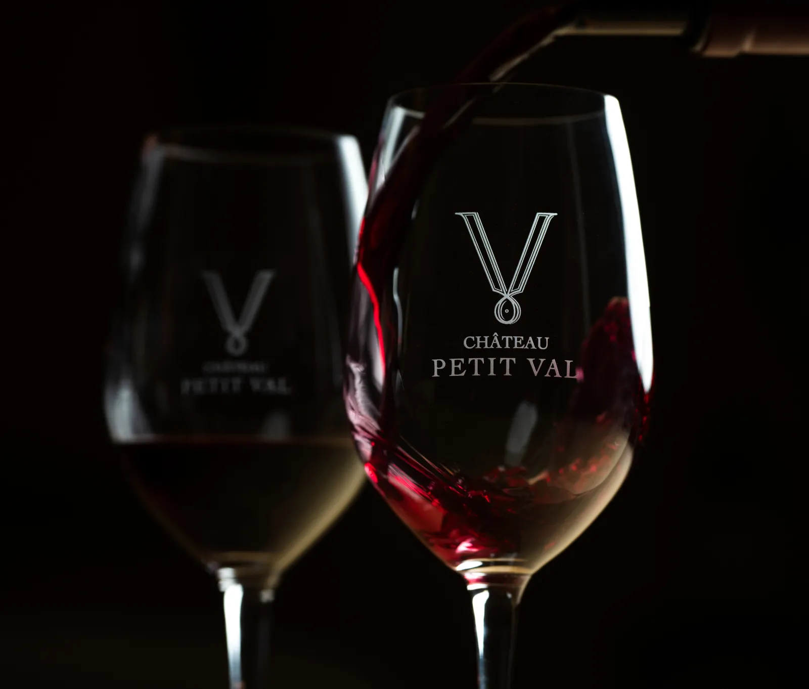 Deux verres de vin gravés du logo Château Petit Val, l'un contenant du vin rouge et l'autre du vin blanc, sur fond noir.