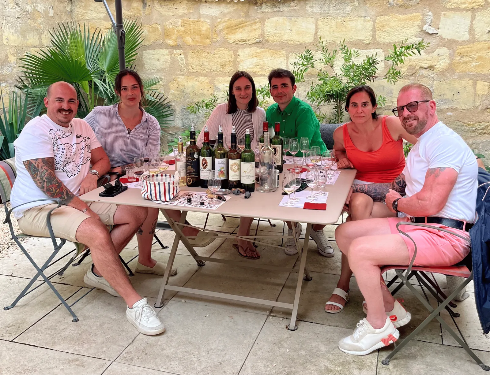 Six amis assis autour d'une table de dégustation avec plusieurs bouteilles de vin, dans une cour pavée devant un mur en pierre à Bordeaux