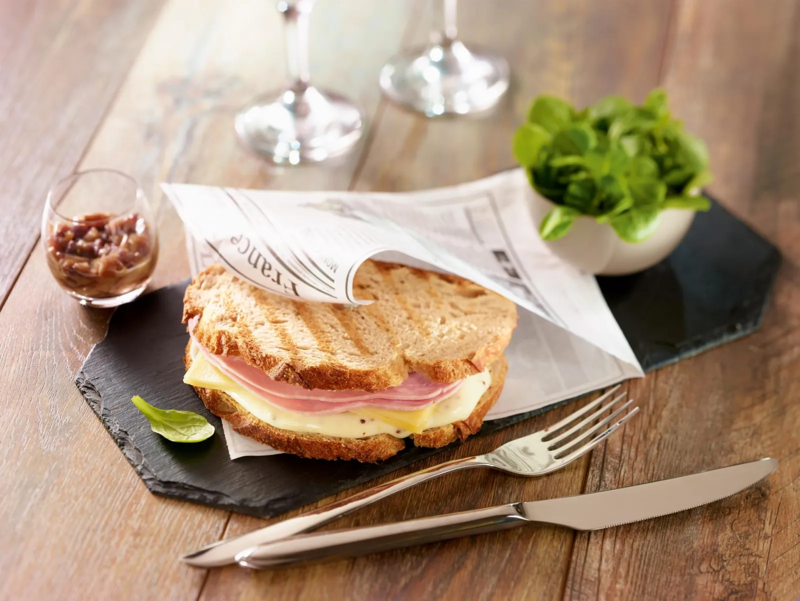 Sandwich jambon-fromage servi sur ardoise noire avec salade fraîche, verre de boisson, couverts et journal à l'Ibis Kitchen Lounge de Bordeaux