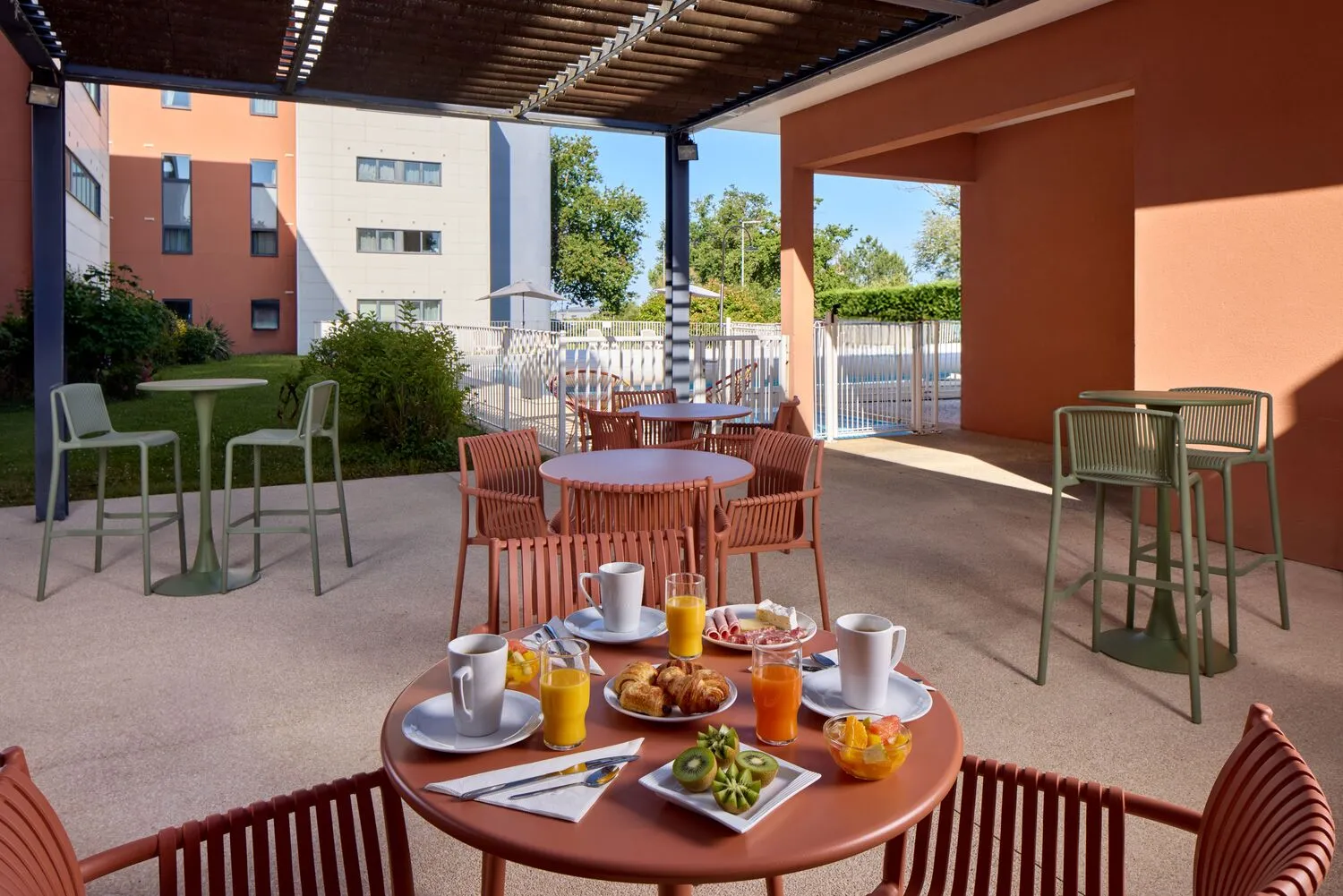 Terrasse couverte d'un hôtel à Bordeaux avec tables de petit-déjeuner, chaises rouges et vertes, café et jus d'orange, bâtiments résidentiels en arrière-plan sous un ciel bleu.