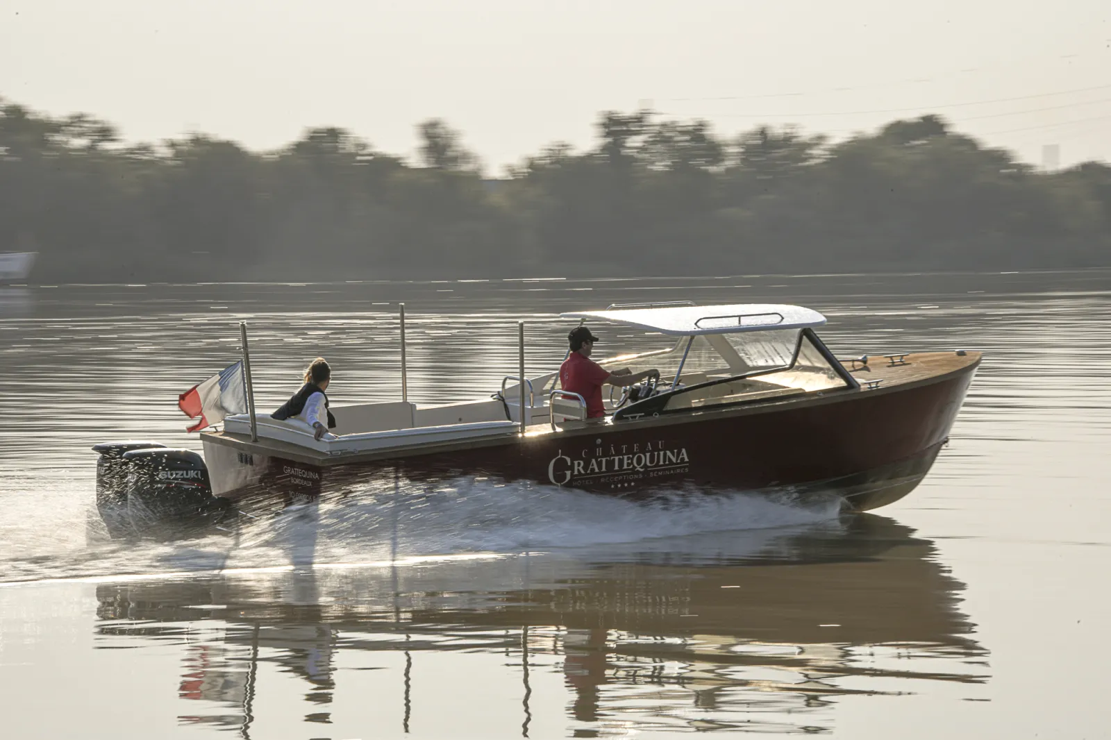 Le bateau-taxi Grattequina amarré sur la Garonne à Bordeaux, avec deux passagers à bord et un opérateur, reflété dans l'eau calme avec la forêt en arrière-plan.