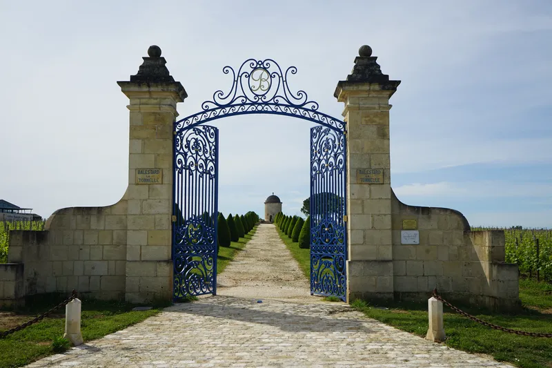 Portail d'entrée orné du Château Balestard La Tonnelle à Bordeaux, composé d'une grille en fer forgé bleu entre deux piliers de pierre, avec une allée de vignes s'étendant vers un bâtiment en arrière-plan