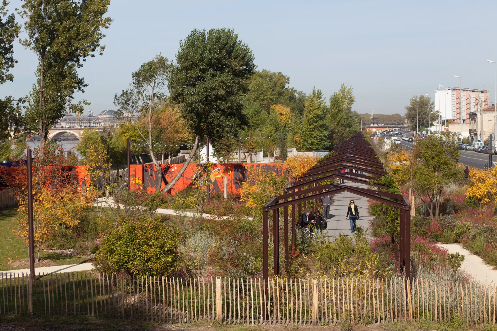 Parc aux Angéliques à Bordeaux en automne, avec une pergola en bois, des arbres aux feuilles colorées, une clôture blanche, et une fresque murale orange en arrière-plan. La Garonne et ses ponts sont visibles à l'horizon.