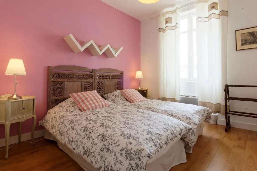 Chambre à coucher élégante avec mur rose, lit double à tête de lit sculptée, draps à motifs floraux, coussins rayés roses et blancs, lampes de chevet, parquet, rideaux blancs et décoration murale géométrique.