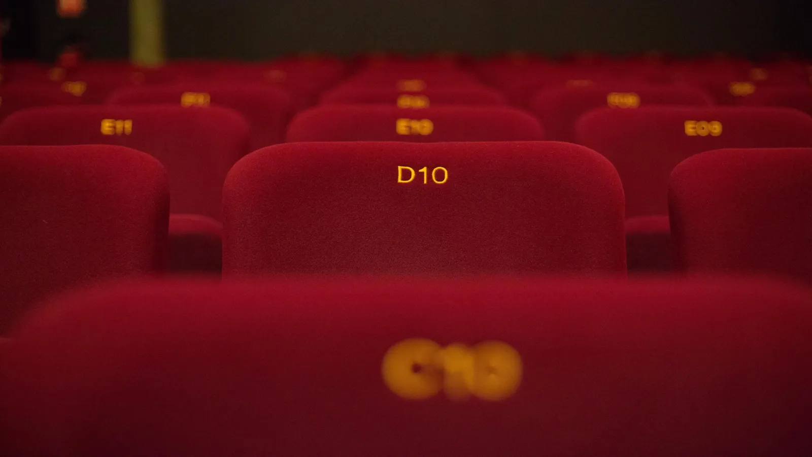 Sièges rouges vides d'une salle de cinéma avec le siège D10 visible au centre et des numéros de places en jaune. Deux tasses de boisson sont posées sur un accoudoir au premier plan.