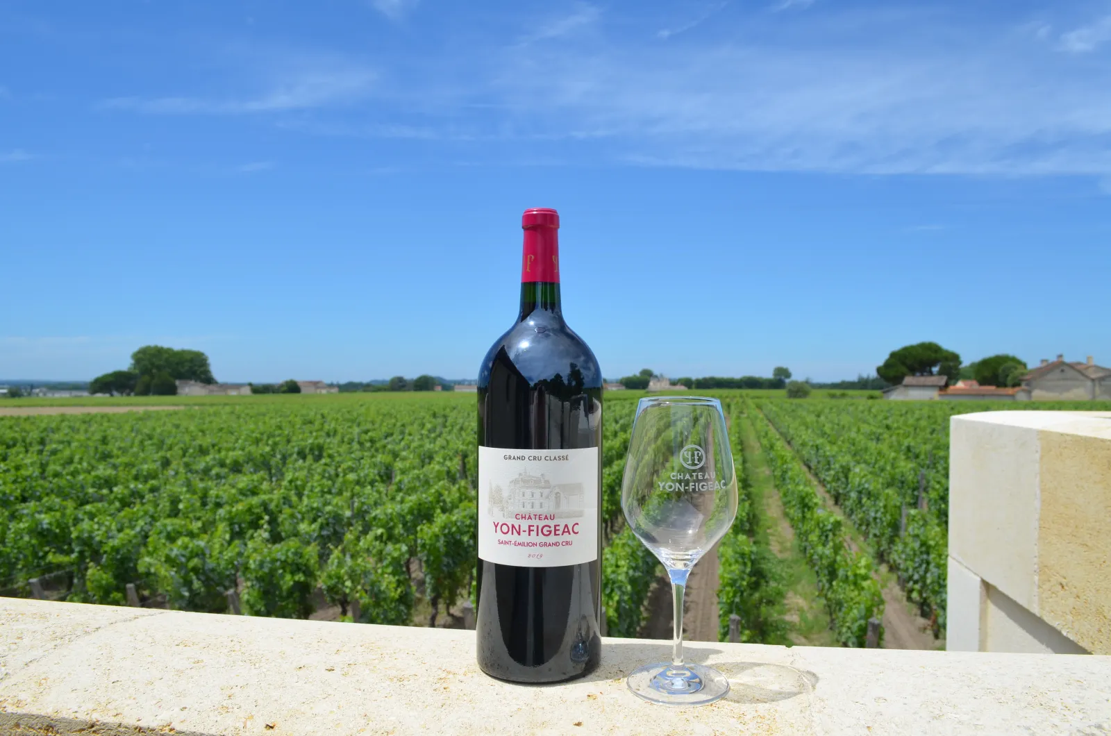 Bouteille de vin rouge Château Yon-Figeac Grand Cru Classé de Bordeaux avec un verre gravé du château, posée sur un muret de pierre face à des vignobles verdoyants sous un ciel bleu.