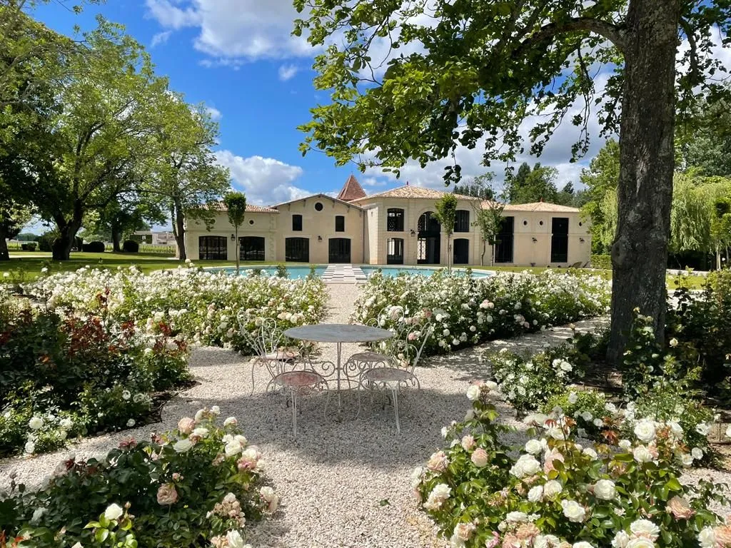 Vue frontale du Château Montlabert à Bordeaux, façade beige avec tour centrale, entouré d'un jardin avec roses blanches et table de fer forgé