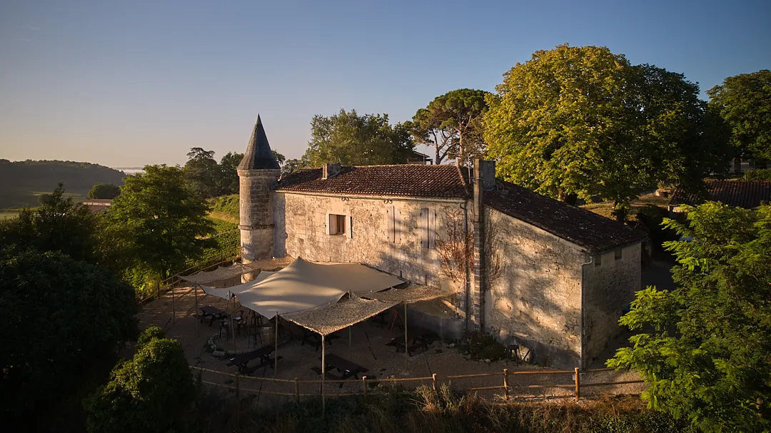 Vue aérienne du Château Tour Baladoz, un manoir de pierre avec une tour conique pointue, entouré de verdure luxuriante et d'arbres, avec une terrasse couverte en premier plan à Bordeaux.