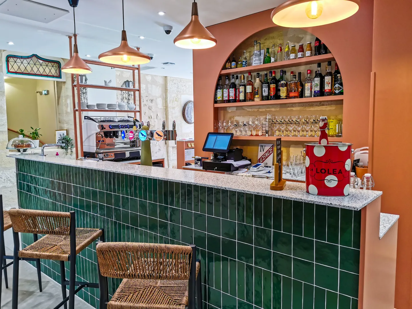 Comptoir de bar moderne avec carrelage vert, étagère murale en alcôve remplie de bouteilles d'alcool et de verres, machine à espresso, tabourets en rotin, luminaires suspendus en cuivre et murs orange dans un café-bar de Bordeaux.